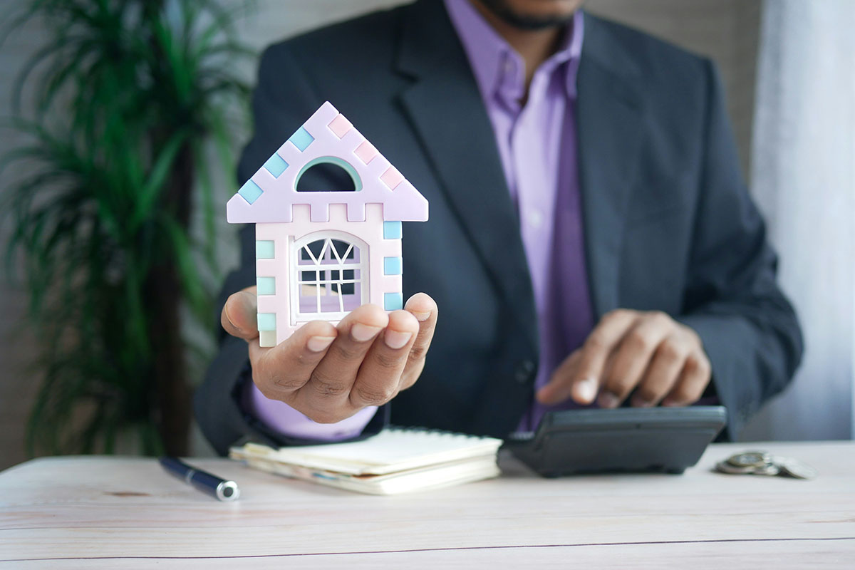A business professional in a suit holds a colorful toy house in one hand while operating a calculator with the other, surrounded by a notepad and a pen on a wooden desk.