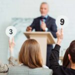 A blurred auctioneer stands at a podium while audience members raise paddles displaying the numbers 3 and 9.