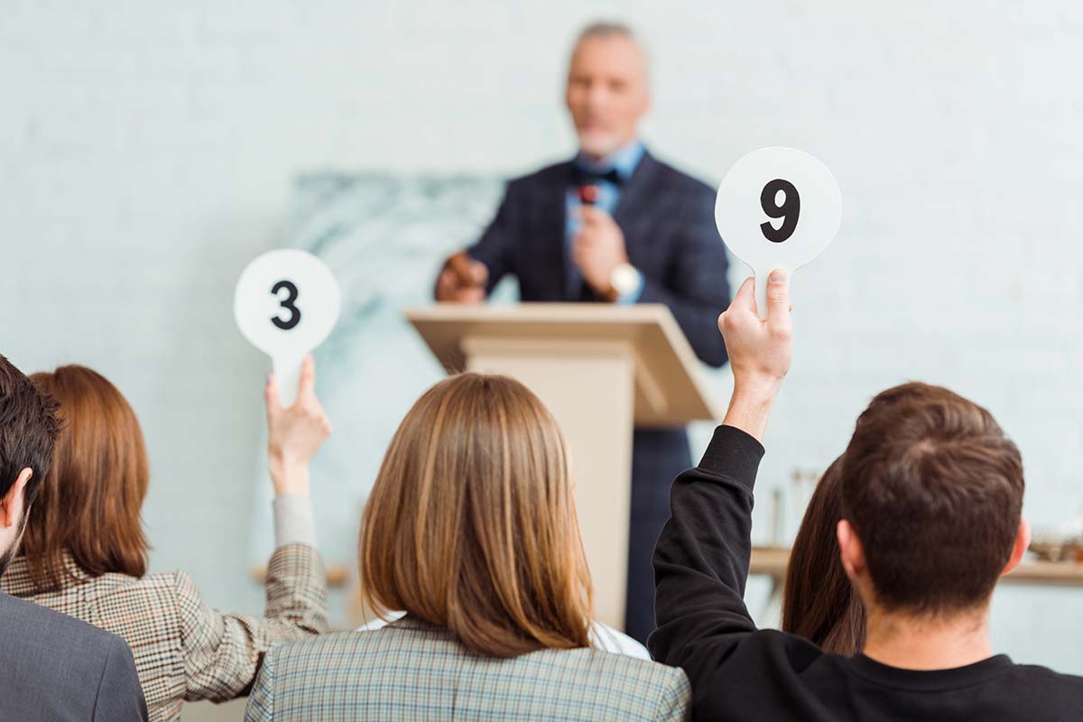 A blurred auctioneer stands at a podium while audience members raise paddles displaying the numbers 3 and 9.