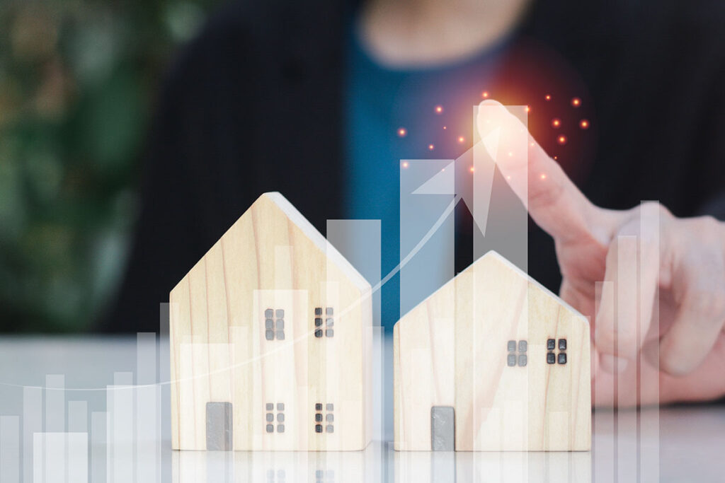 A person's hand points at two wooden house models with a rising graph and glowing elements in the background, representing growth in real estate or housing market trends.