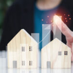 A person's hand points at two wooden house models with a rising graph and glowing elements in the background, representing growth in real estate or housing market trends.