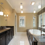A stylish bathroom featuring a dark wood vanity with two sinks, a large white soaking tub, and marble accents. The walls are painted a soft beige, complemented by lighting fixtures and large windows that provide natural light. The floor is adorned with polished tiles in a mosaic pattern.