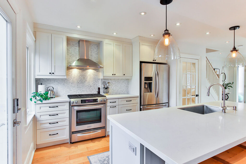 A modern kitchen featuring white cabinetry, a stainless steel oven, and a sleek island with a sink. The space is well-lit with pendant lighting and recessed lights, showcasing a light-colored backsplash and warm wooden flooring.