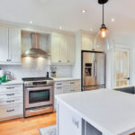 A modern kitchen featuring white cabinetry, a stainless steel oven, and a sleek island with a sink. The space is well-lit with pendant lighting and recessed lights, showcasing a light-colored backsplash and warm wooden flooring.