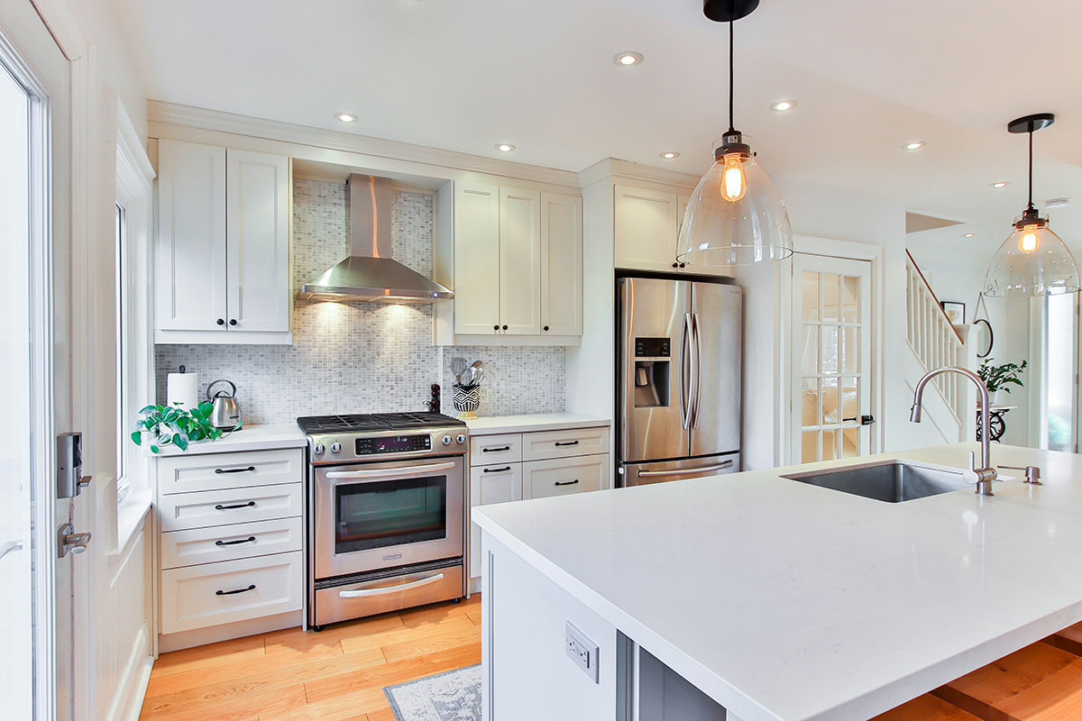 A modern kitchen featuring white cabinetry, a stainless steel oven, and a sleek island with a sink. The space is well-lit with pendant lighting and recessed lights, showcasing a light-colored backsplash and warm wooden flooring.