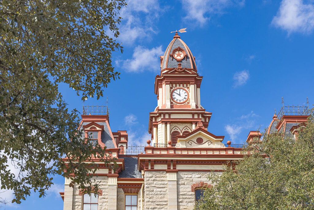 The historic Second Empire style Caldwell County Courthouse tower in Lockhart, Texas, framed by leafy green tree branches against a bright blue sky.