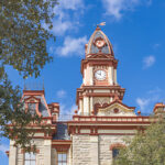 The historic Second Empire style Caldwell County Courthouse tower in Lockhart, Texas, framed by leafy green tree branches against a bright blue sky.