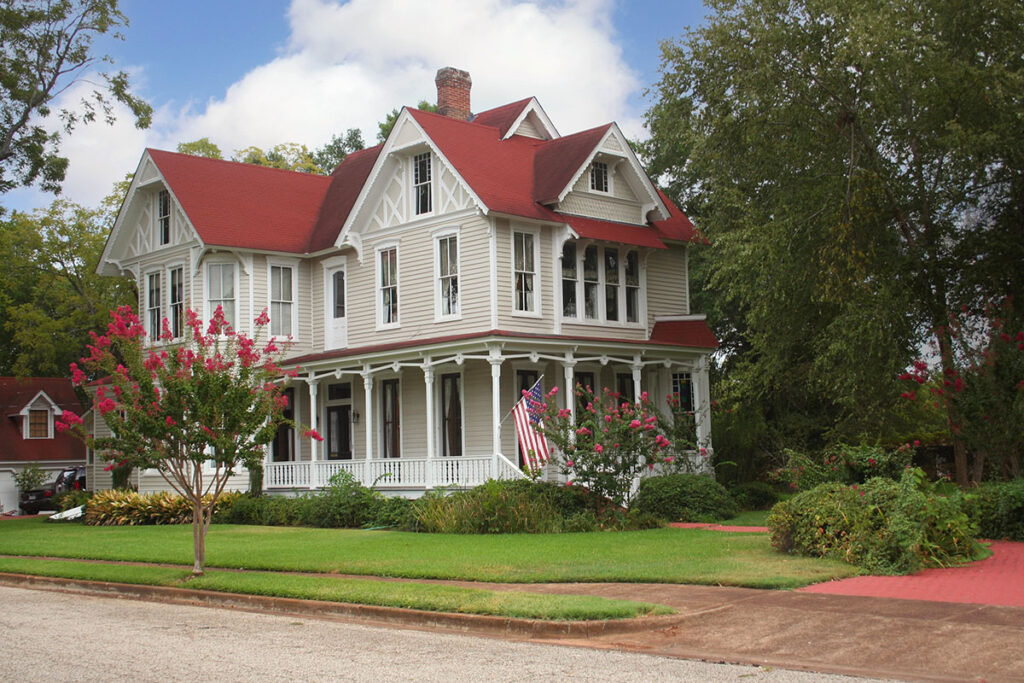 A historic two-story Queen Anne Victorian house with light beige siding, a red gabled roof, and a wrap-around front porch under a sunny blue sky.