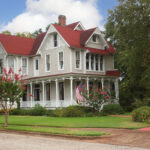 A historic two-story Queen Anne Victorian house with light beige siding, a red gabled roof, and a wrap-around front porch under a sunny blue sky.