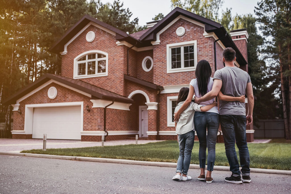 Rear view of a family standing with their arms around each other, looking at a large two-story red brick suburban home.