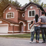 Rear view of a family standing with their arms around each other, looking at a large two-story red brick suburban home.