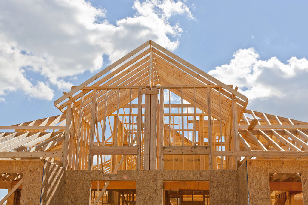 A low-angle view of a wood-frame house under construction, showing structural wall studs and roof trusses against a partly cloudy blue sky.