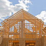 A low-angle view of a wood-frame house under construction, showing structural wall studs and roof trusses against a partly cloudy blue sky.