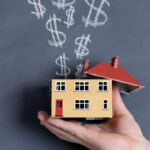A hand holding a small yellow model house with its red roof opened, as chalk-drawn dollar signs rise like smoke against a dark slate background.
