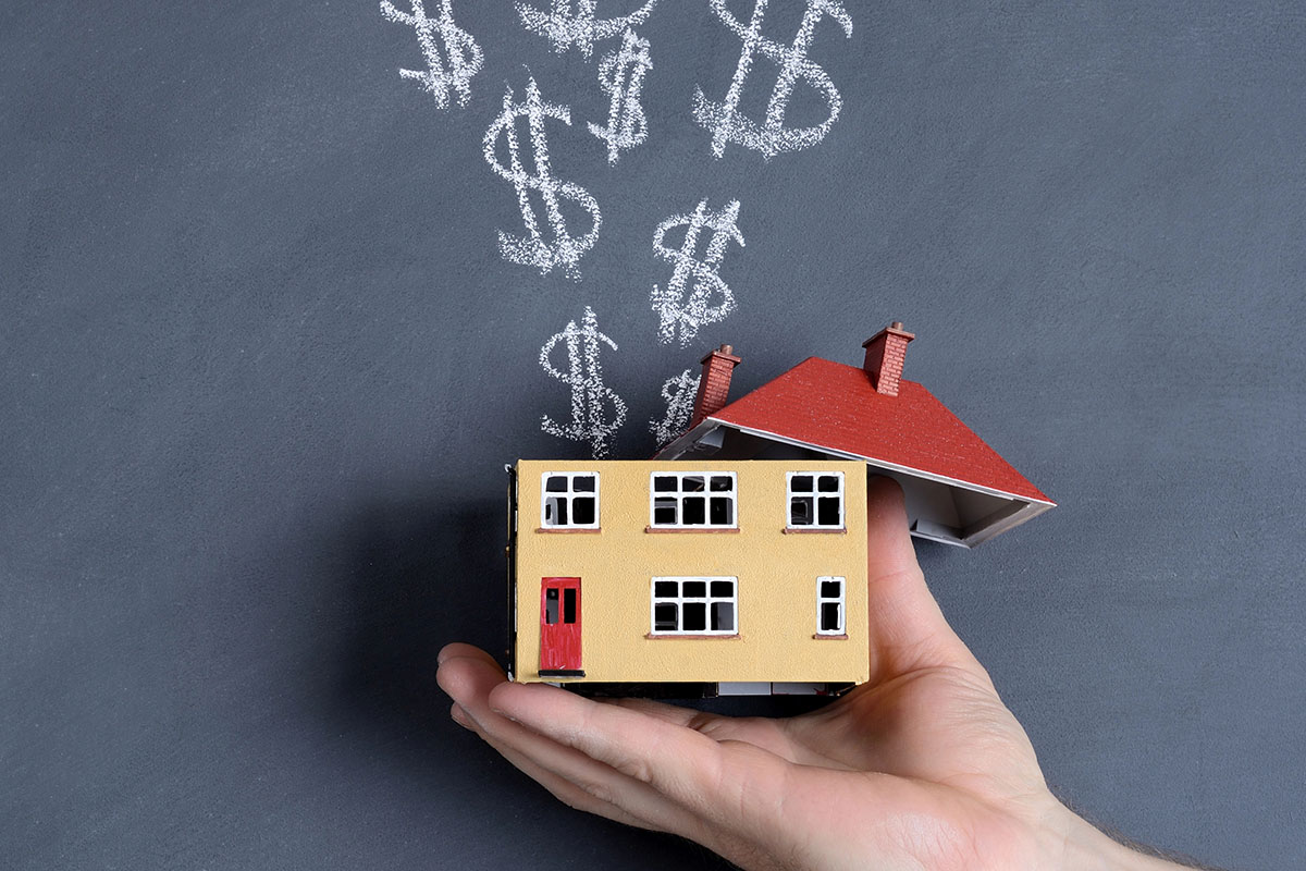 A hand holding a small yellow model house with its red roof opened, as chalk-drawn dollar signs rise like smoke against a dark slate background.