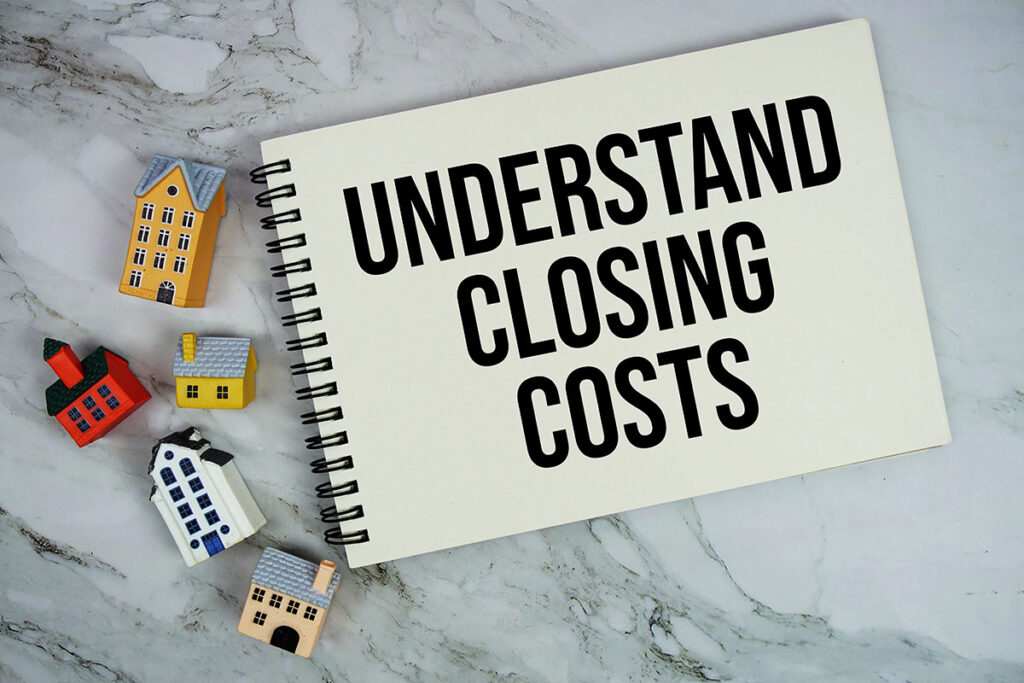 A top-down view of an open spiral notebook with the text "UNDERSTAND CLOSING COSTS" printed on the page, surrounded by miniature colorful houses on a white marble tabletop.
