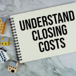 A top-down view of an open spiral notebook with the text "UNDERSTAND CLOSING COSTS" printed on the page, surrounded by miniature colorful houses on a white marble tabletop.